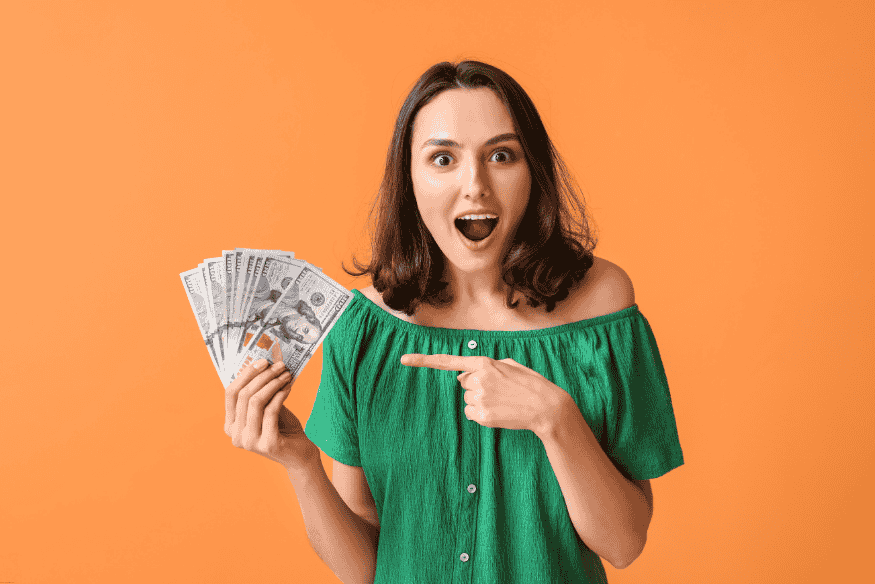 Mujer joven sorprendida sosteniendo billetes de dólar en fondo naranja.