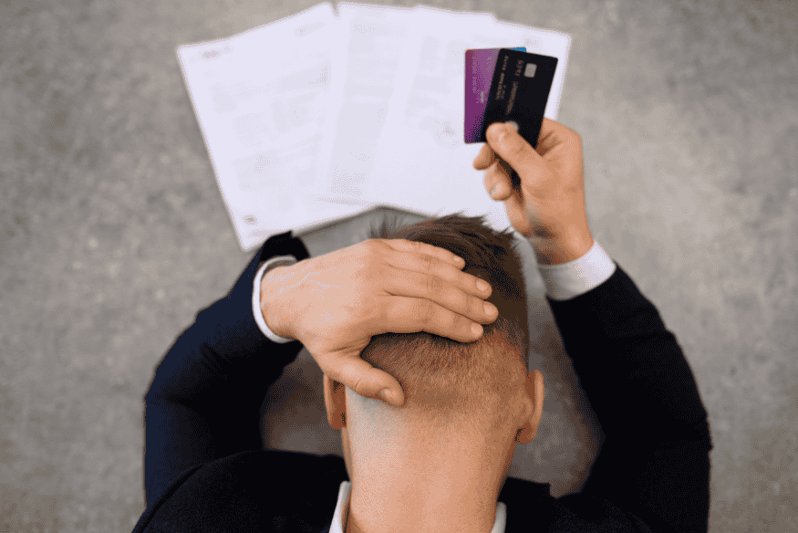 Hombre con expresión de estrés sujetando tarjetas de crédito y revisando documentos financieros.