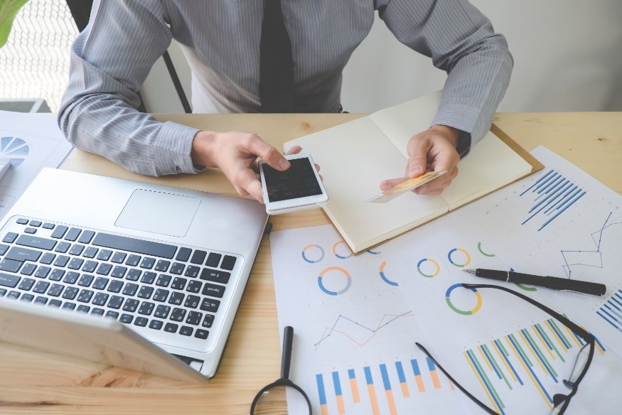 Persona en escritorio con computadora portátil, teléfono y documentos financieros con gráficos.