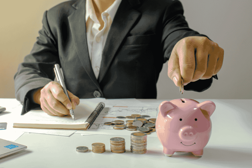 Persona de negocios escribiendo mientras deposita monedas en una alcancía rosada.