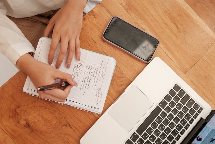 Manos escribiendo en un cuaderno con smartphone y laptop sobre la mesa.