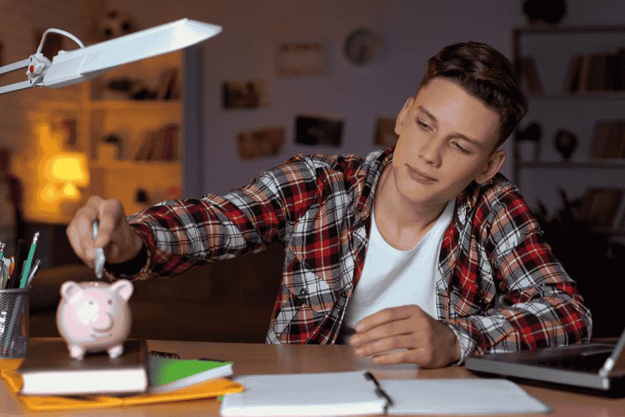 Joven depositando una moneda en una alcancía rosa en su escritorio de estudio.