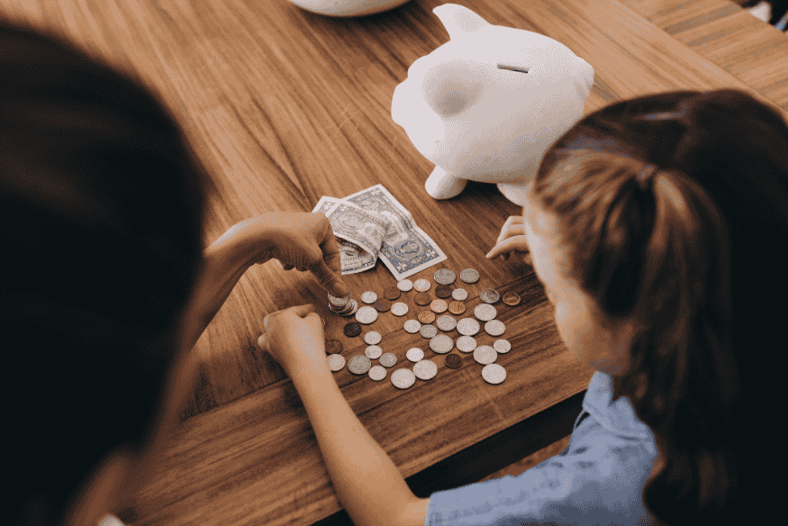Niño y adulto contando monedas y billetes en una mesa junto a una alcancía blanca.