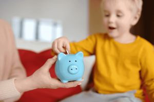 Niño pequeño insertando una moneda en una alcancía azul sostenida por un adulto.
