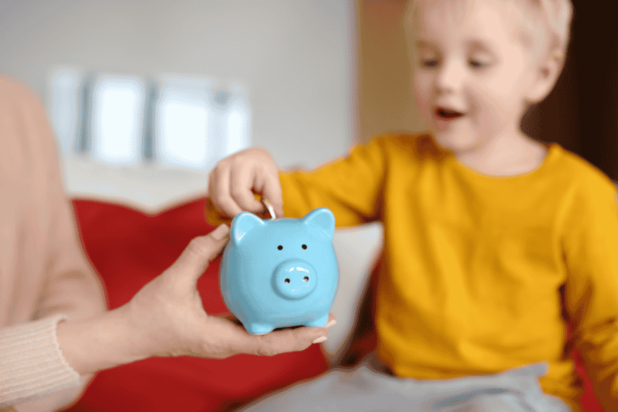 Niño pequeño insertando una moneda en una alcancía azul sostenida por un adulto.