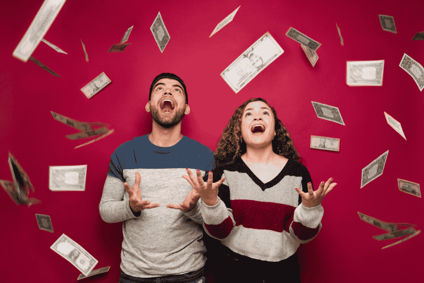 Pareja joven celebrando mientras billetes caen sobre ellos con fondo rojo.