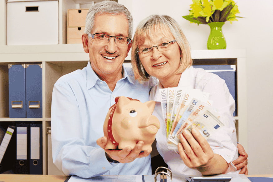 Pareja mayor sosteniendo una alcancía y billetes de euro con actitud alegre.