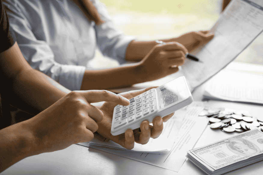 Manos de una pareja usando una calculadora y revisando documentos financieros con billetes y monedas sobre la mesa.