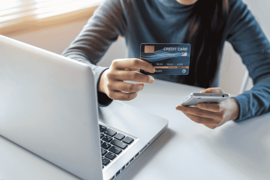 Persona con tarjeta de crédito y celular, realizando consulta o compra online desde laptop.