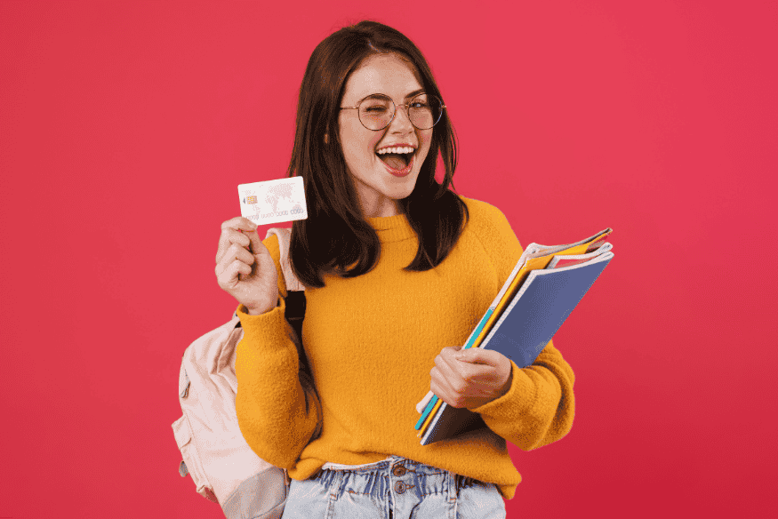 Estudiante emocionada mostrando su tarjeta de crédito con fondo rosa y útiles escolares.