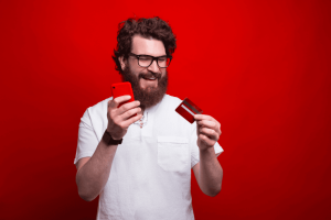 Hombre feliz con tarjeta de crédito y smartphone en fondo rojo